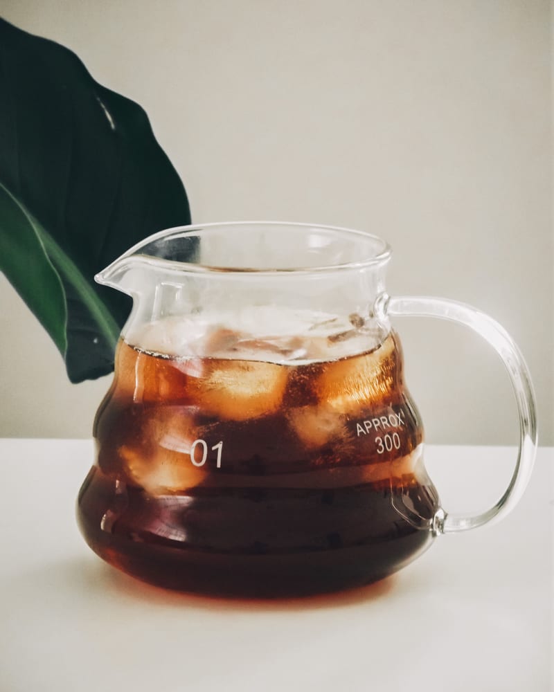A glass carafe of cold brew over ice, beside a large green leaf.