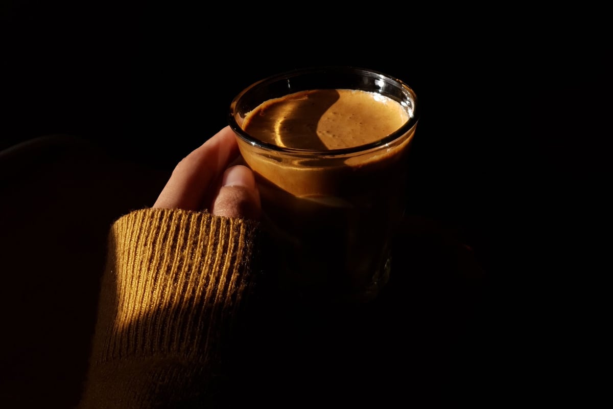 A glass of espresso held in one hand, lit by a single shaft of light.
