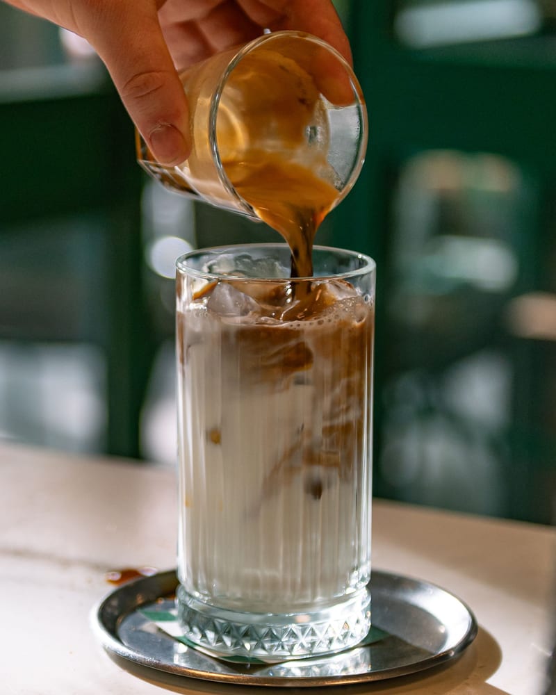 Espresso poured from a small glass into a tall glass of milk over ice.