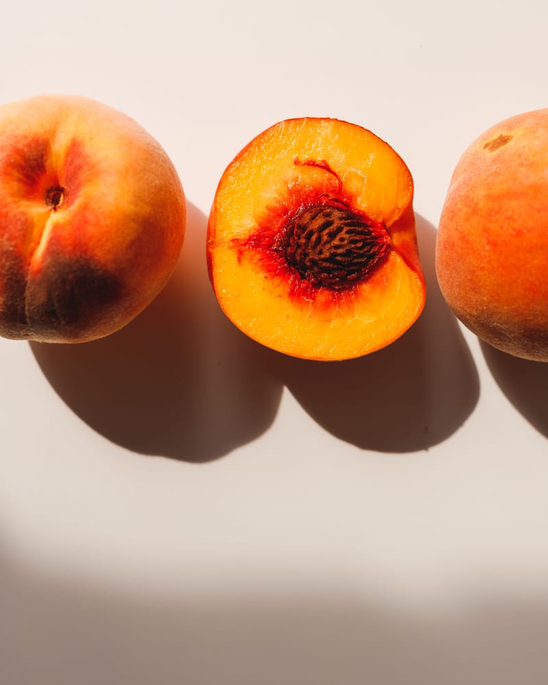 Three ripe peaches on a pale background, the middle one halved to show the stone.
